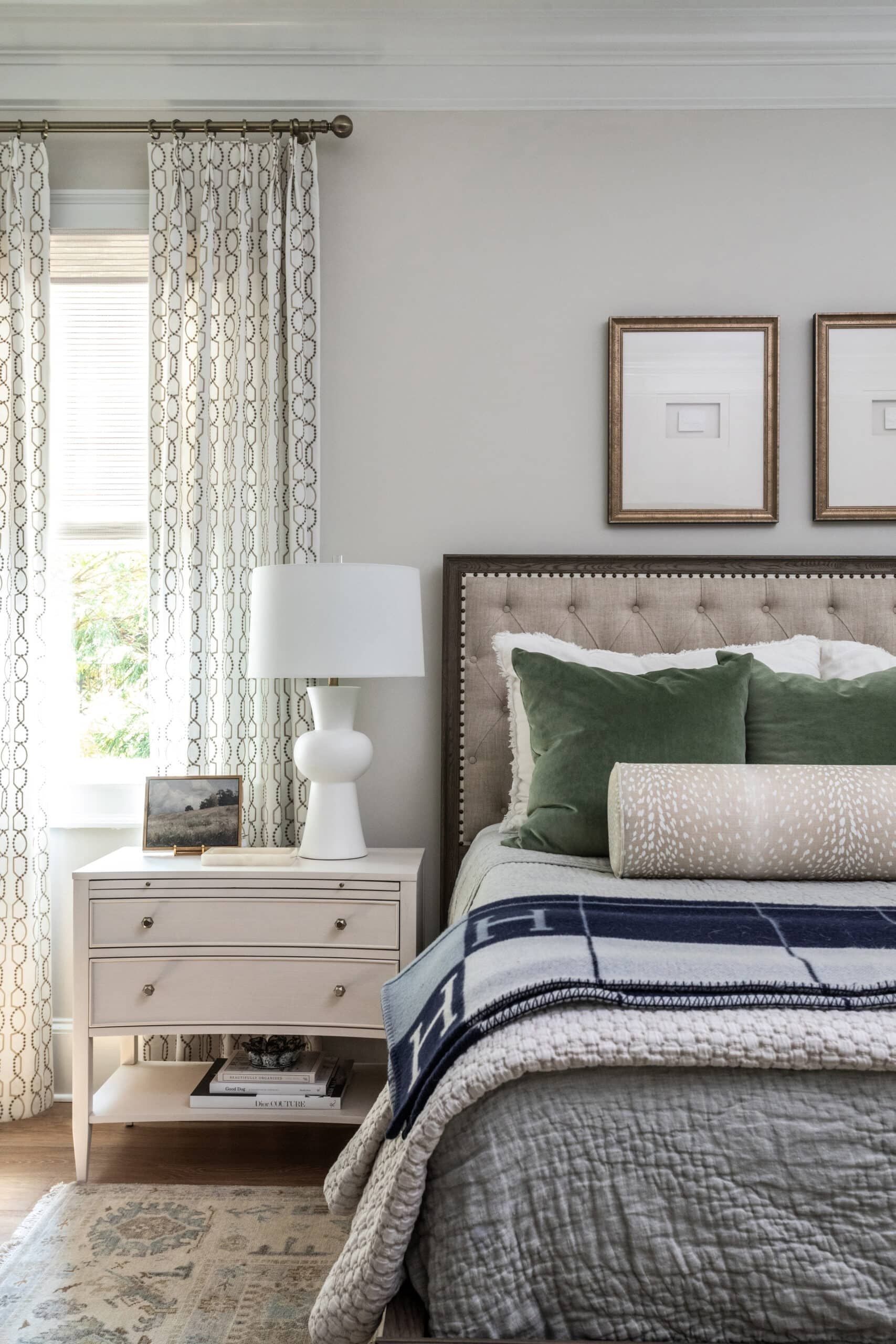 Cream curtains with a dark trellis-and-dot pattern, layered over a woven shade beside a window in a bedroom with a tufted headboard.